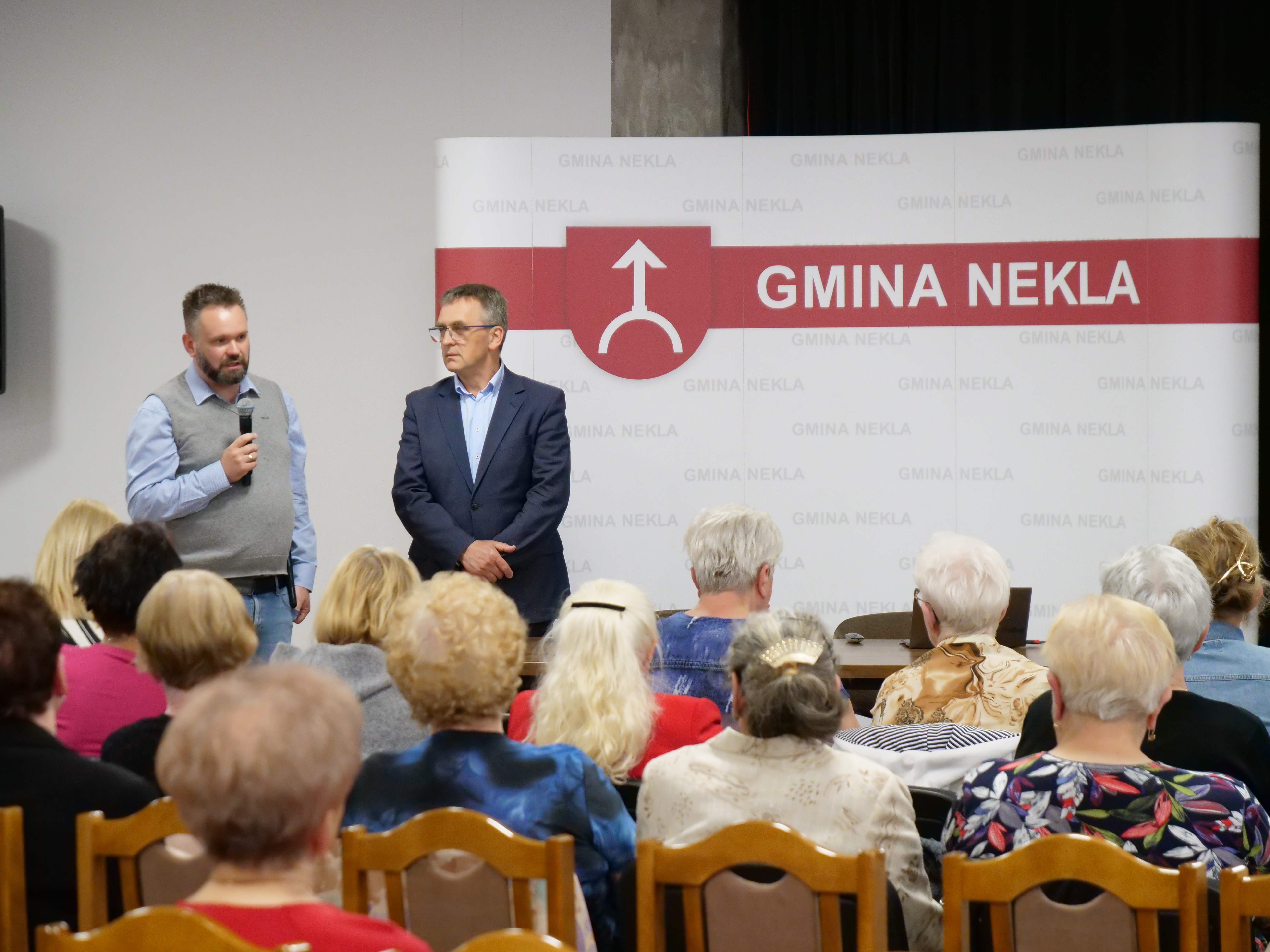 Dwóch mężczyzn przemawia do grupy osób, siedzącej na krzesłach. Jeden trzyma mikrofon. W tle baner z napisem Gmina Nekla.